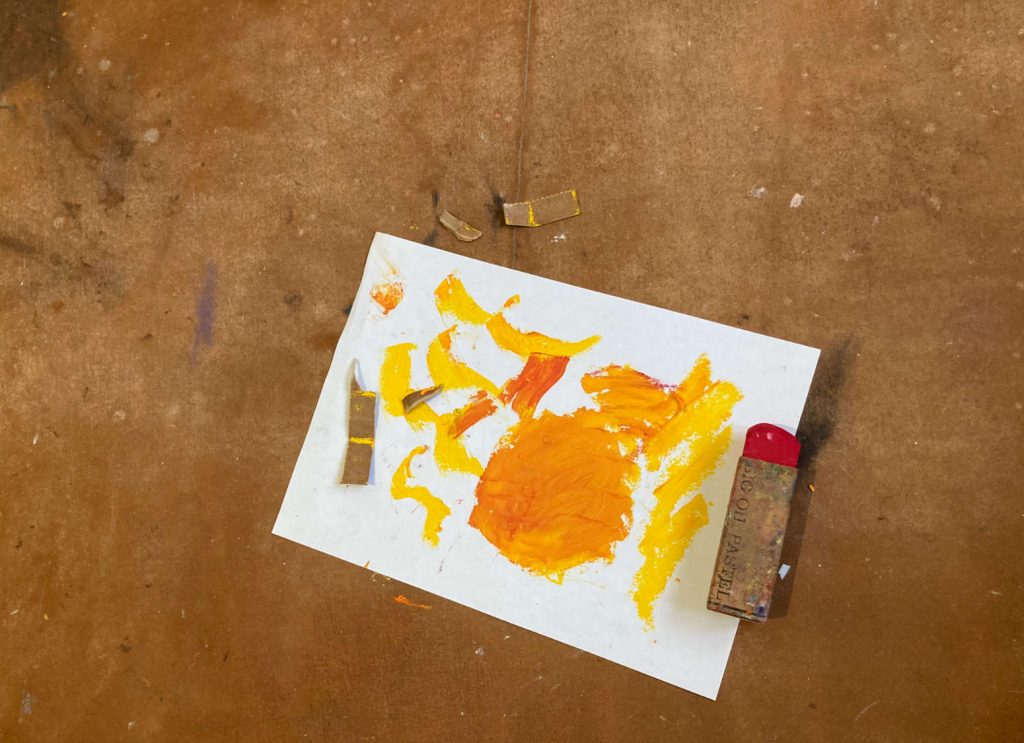 a white sheet of paper with orange test colors next to a large red oil pastel stick